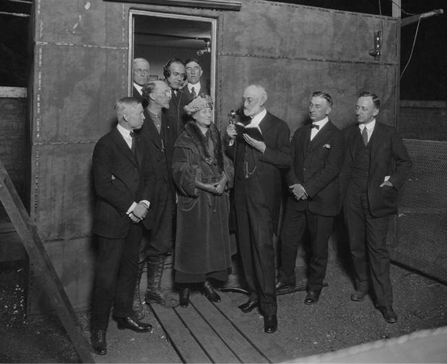 Black-and-white photograph of Heber J. Grant speaking into a microphone, surrounded by men and a woman in formal attire, inside an industrial setting.