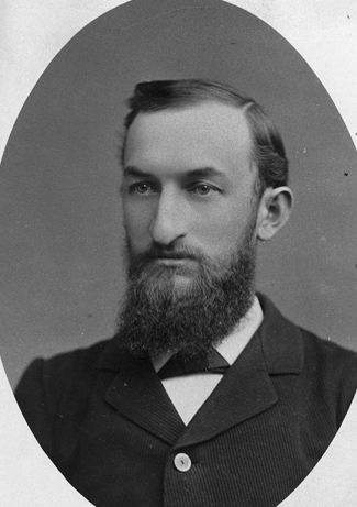 Black-and-white studio portrait of a young man with a full beard, wearing a dark suit and bow tie, shown from the chest up.