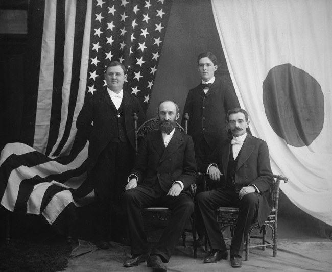 Black-and-white group portrait of four men in formal suits, two seated and two standing, posed indoors with large American and Japanese flags behind them.