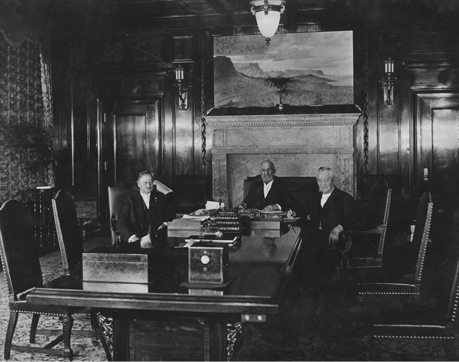 Black-and-white photograph of three men seated around a desk in a formal office interior, surrounded by dark wood furnishings