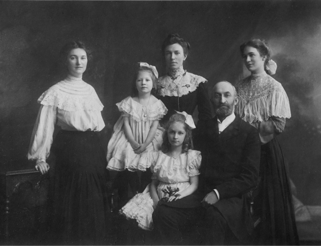 Black-and-white family portrait showing a seated man with a woman and four girls standing and seated around him, all dressed in early twentieth-century clothing