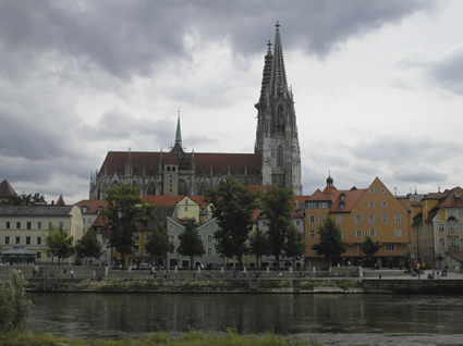 Photograph of the Regensburg Cathedral rising above riverside buildings along the Danube River, with the city skyline reflected in the water.