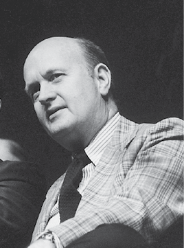 Black-and-white portrait of a middle-aged man in a suit and tie, shown from the chest up against a dark background.