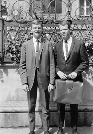 Black-and-white photograph of two young men in suits standing outdoors near a decorative public fountain.